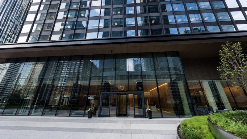 Street-level view of 10 S. Riverside Plaza in Chicago, showing the modern glass office tower entrance with reflective windows, revolving doors, and landscaped planters in the West Loop business district.